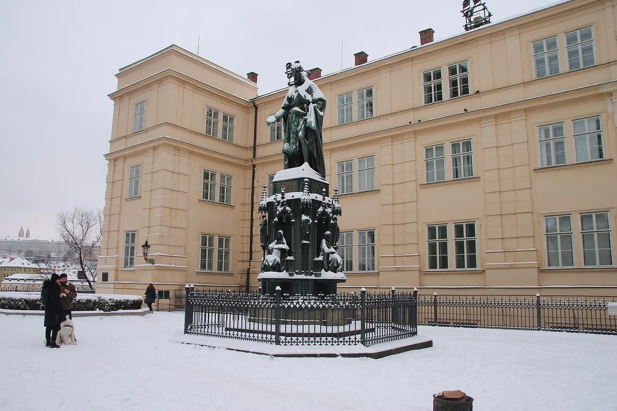 A statue of Charles IV on a stone plinth in a snow-covered square, with a long pale-yellow building behind it
