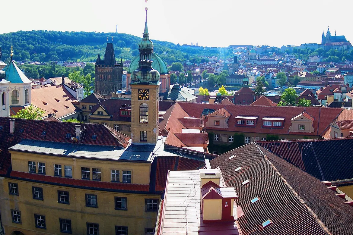 Red roofs of the Old Town seen from the Klementinum's Astronomical Tower, with a church tower and green hills beyond