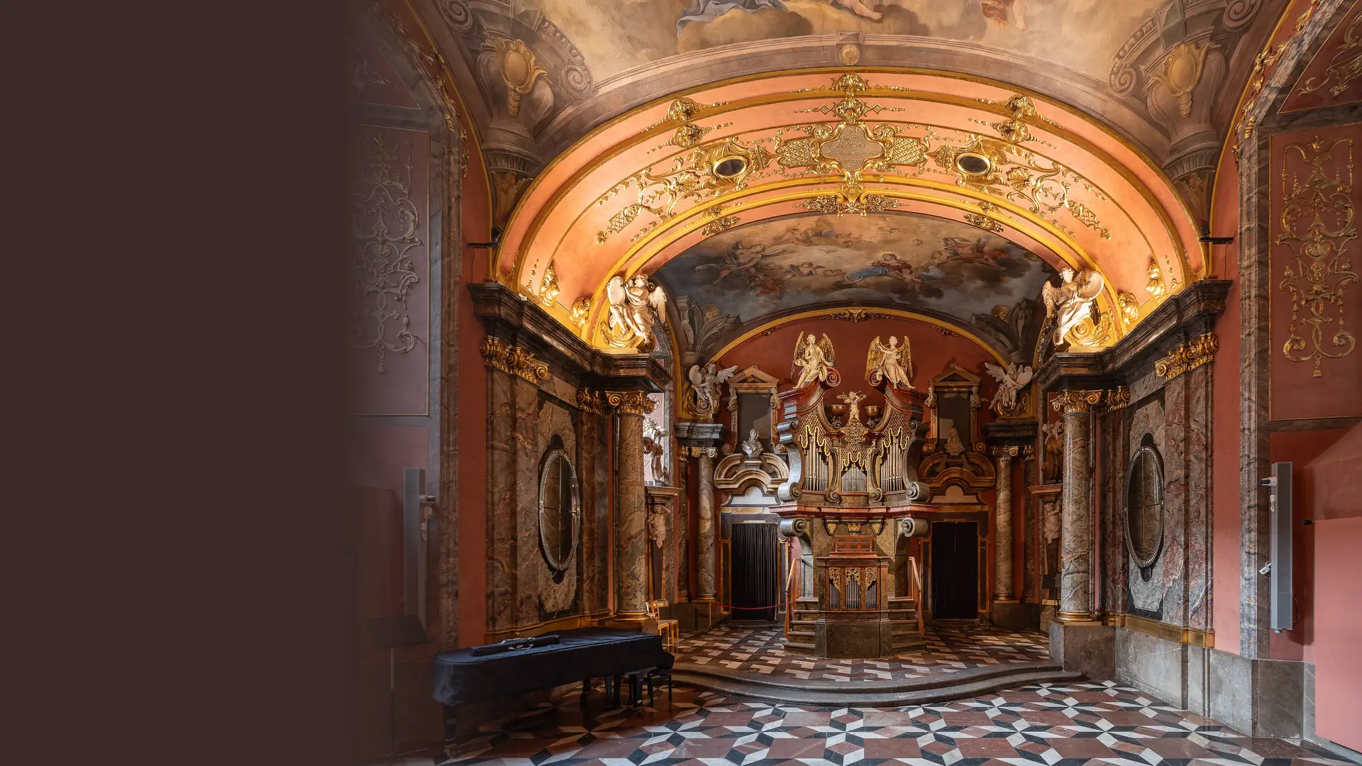 The Mirror Chapel in Prague's Klementinum: a gilded Baroque vault with ceiling frescoes, white angels at the corners and an organ above a marble altar, with a grand piano at the left.